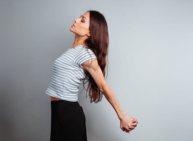 5 bài tập giãn cơ đốt cháy mỡ thừa nhiều hơn 30 phút đi bộ- Ảnh 4. Người phụ nữ xinh đẹp, khỏe khoắn trong trang phục thường ngày đang thực hiện bài tập kéo giãn ngực và vai, nắm chặt tay sau lưng để thư giãn cơ thể trên nền studio màu xanh. Lối sống lành mạnh