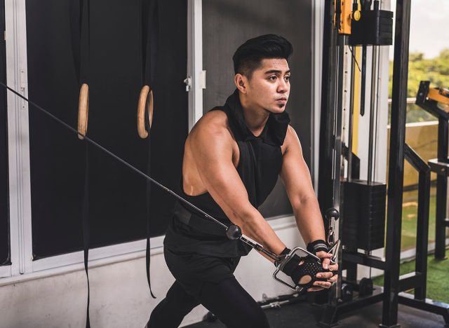 5 bài tập ngực giúp tăng cơ nhiều hơn chống đẩy sau 50 tuổi- Ảnh 5. Một chàng trai châu Á ngoài 20 tuổi, khỏe mạnh và rám nắng, đang tập một loạt bài tập cáp chéo tại phòng tập. Anh mặc áo ba lỗ khoét cổ thấp màu đen có mũ trùm đầu. Anh đang tập cơ ngực trong phòng tập.