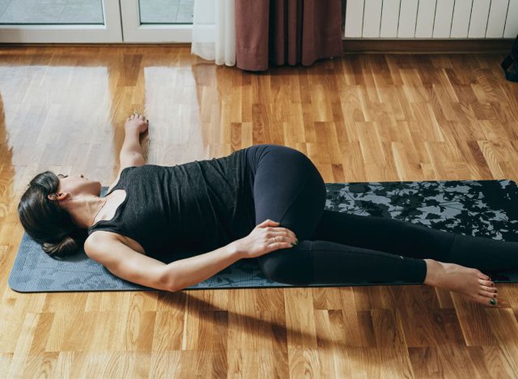 Thực hành tư thế xoay cột sống ngả: Người phụ nữ xinh đẹp mặc đồ thể thao đang thực hiện bài tập xoay cột sống trên tấm thảm tập màu xám trong phòng khách tại nhà
