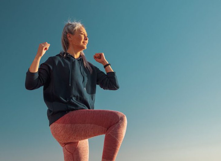 Người phụ nữ năng động mặc quần leggin màu hồng và áo hoodie màu xanh đang tập thể dục, nhấc một đầu gối lên và nhìn về phía trước vào ngày nắng